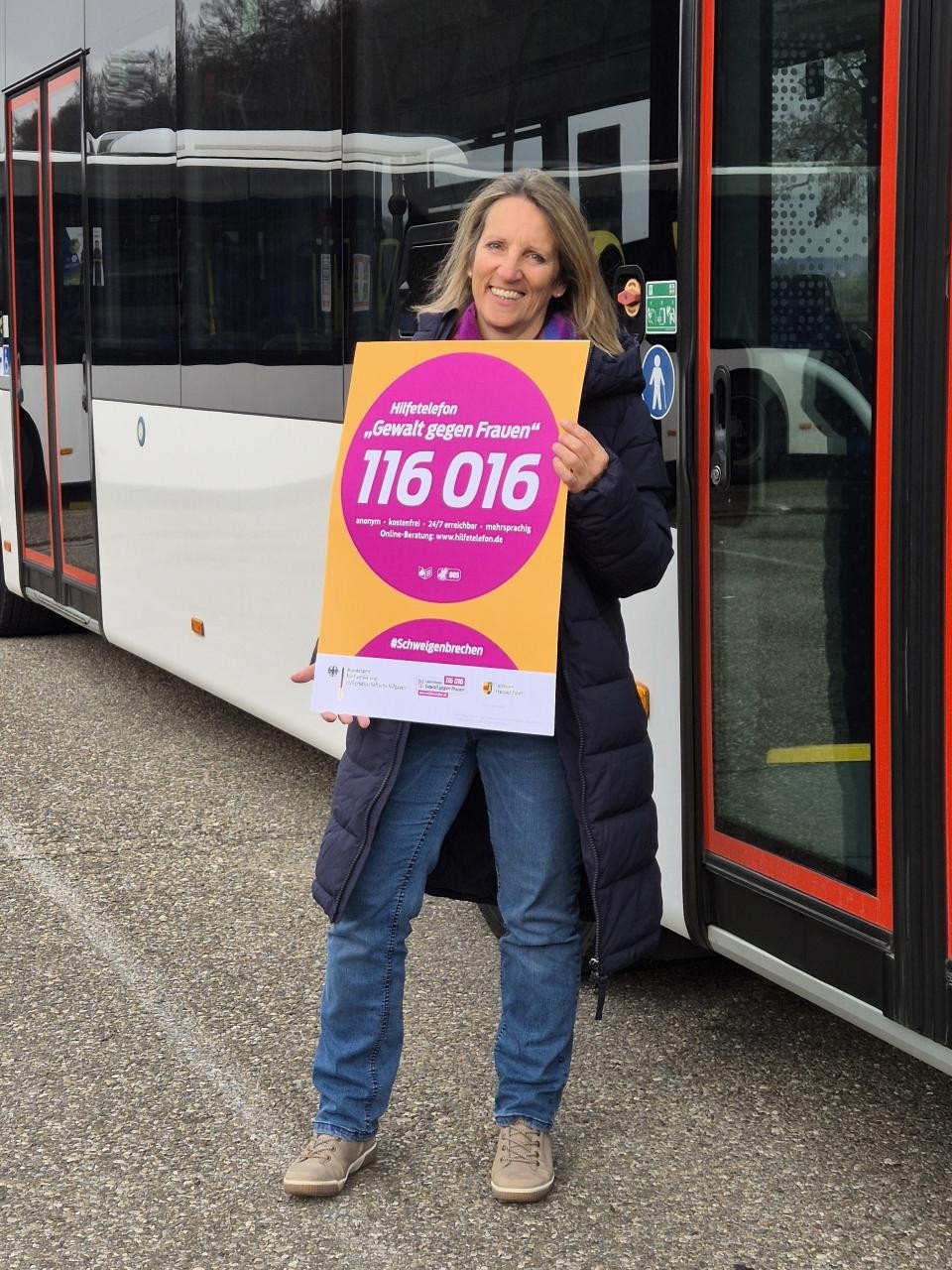 Die Gleichstellungsbeauftragte steht vor einem Bus und hält ein Plakat in die Kamera.