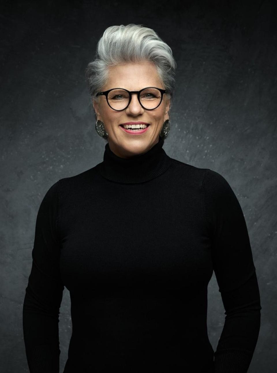 Eine Frau mit schwarzem Pullover, grauen Haaren und Brille lächelt in die Kamera.
