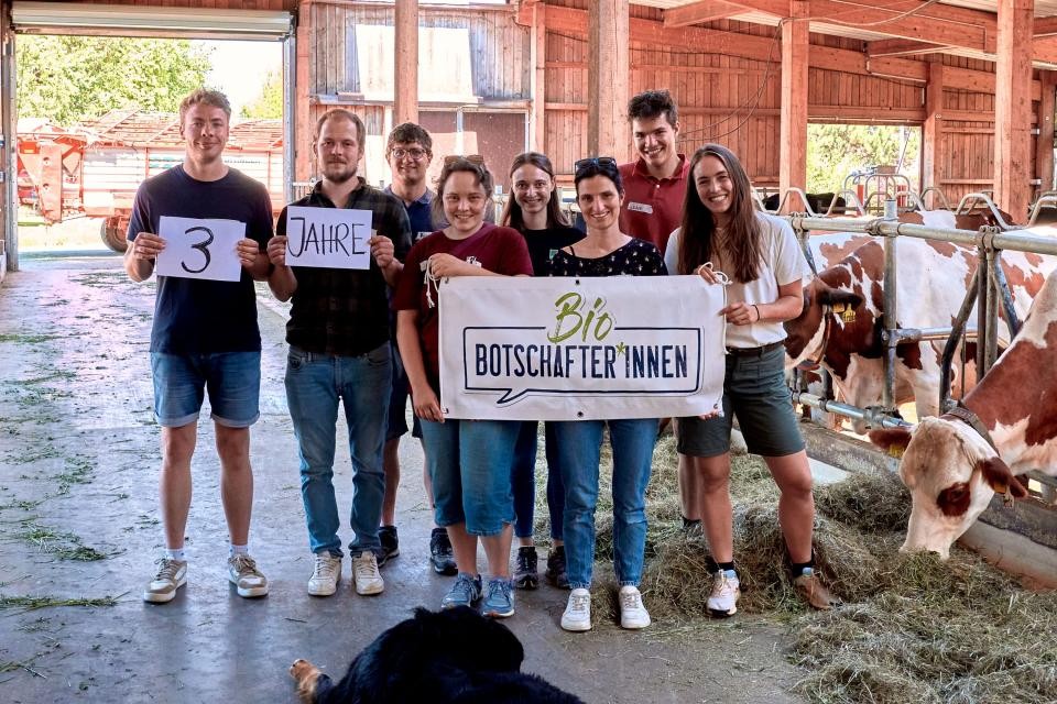 Die Bio-Botschafter stehen in einem Stall umgeben von Kühen und den Schriftzug "3 Jahre Bio-Botschafter" hoch.